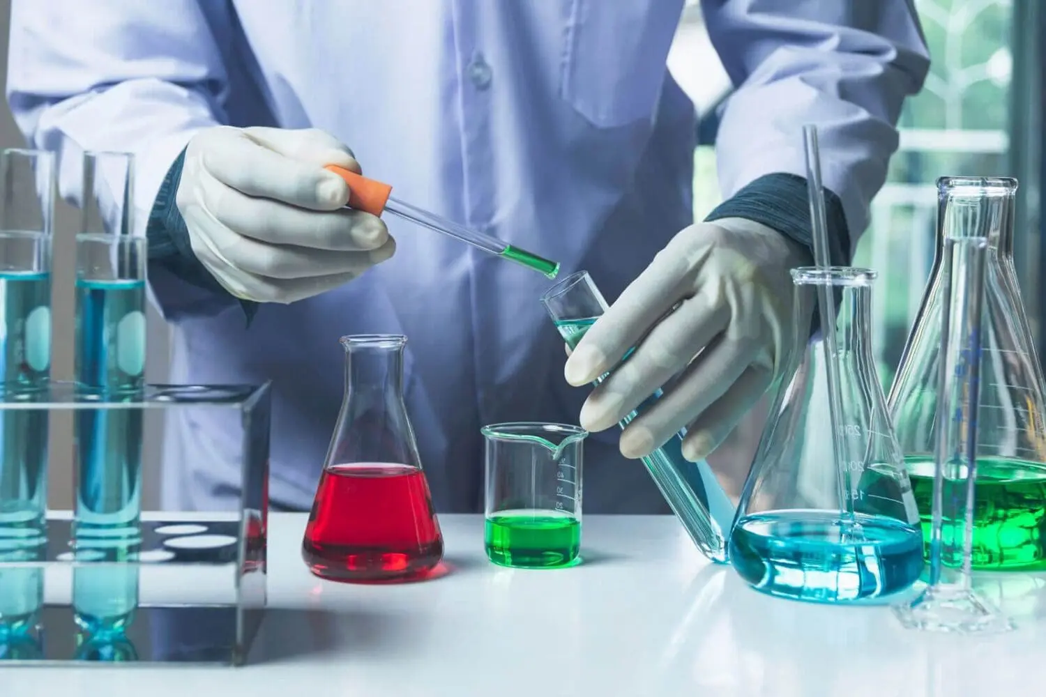 Adherent (formerly Compliance & Risks). A person in a white lab coat and gloves uses a dropper to transfer green liquid into a test tube containing blue liquid, surrounded by various flasks and beakers with colorful solutions on a lab bench.