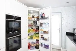 An open, built-in refrigerator filled with food and drinks, next to a black double oven, in a modern white kitchen.
