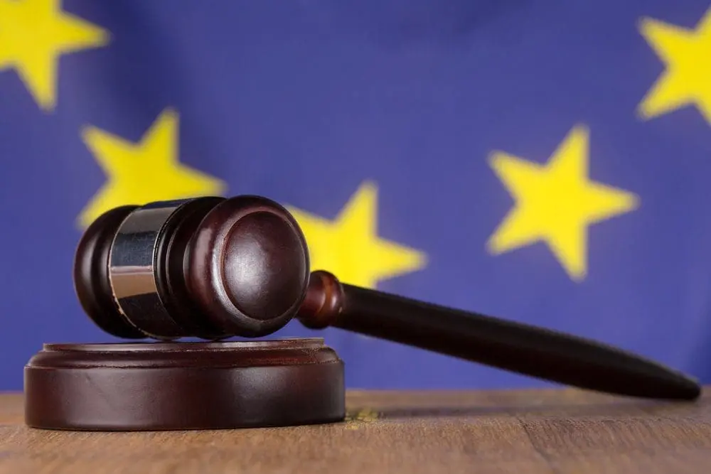 A dark brown wooden gavel on its sound block, with the blurred blue and yellow star European Union flag in the background.