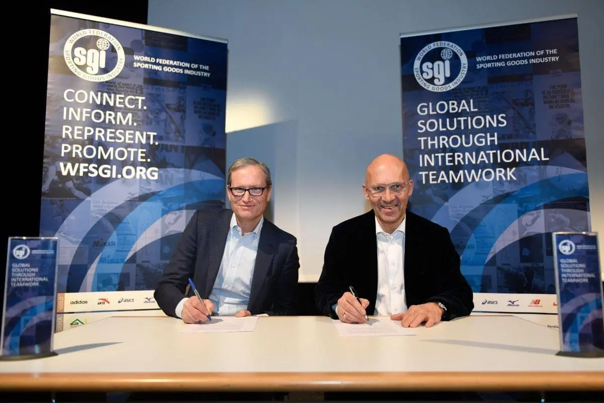 Adherent (formerly Compliance & Risks). Two men, smiling, sign documents at a table with WFSGI banners in the background.