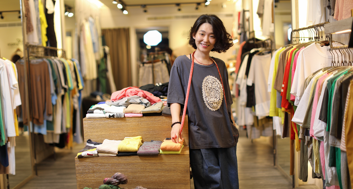 Adherent (formerly Compliance & Risks). A smiling woman with short, dark hair and a measuring tape around her neck stands in a clothing store with racks of colorful clothes.