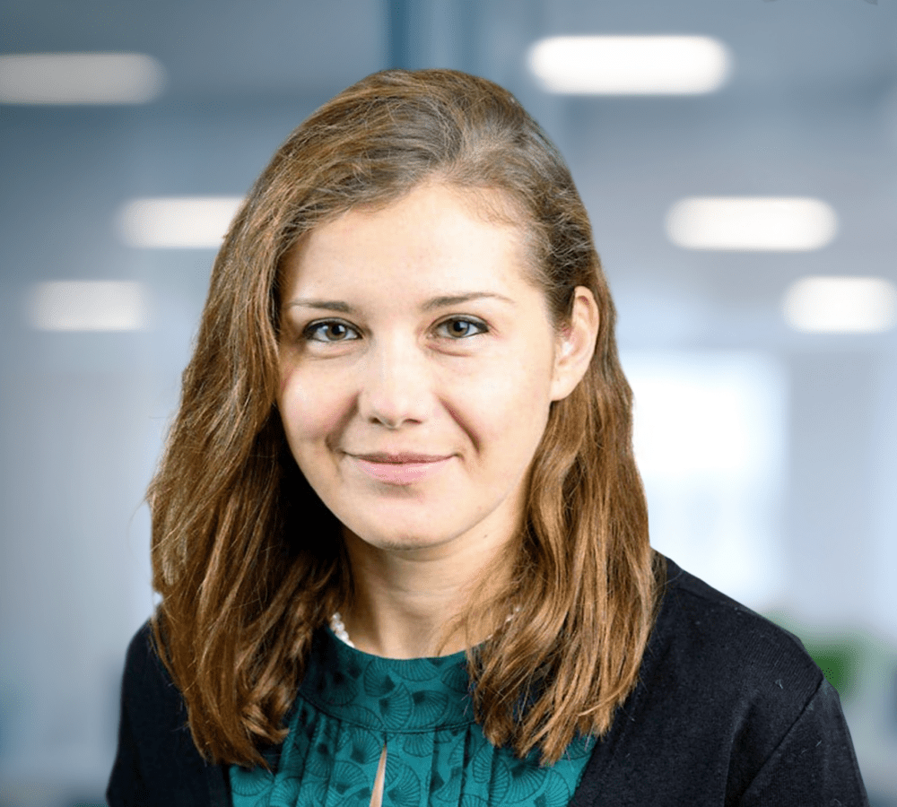 Adherent (formerly Compliance & Risks). Headshot of a smiling woman with brown hair and a green top in a blurred office background.