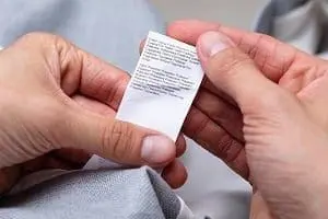 Adherent (formerly Compliance & Risks). Close-up of hands holding a white clothing care label with small text.