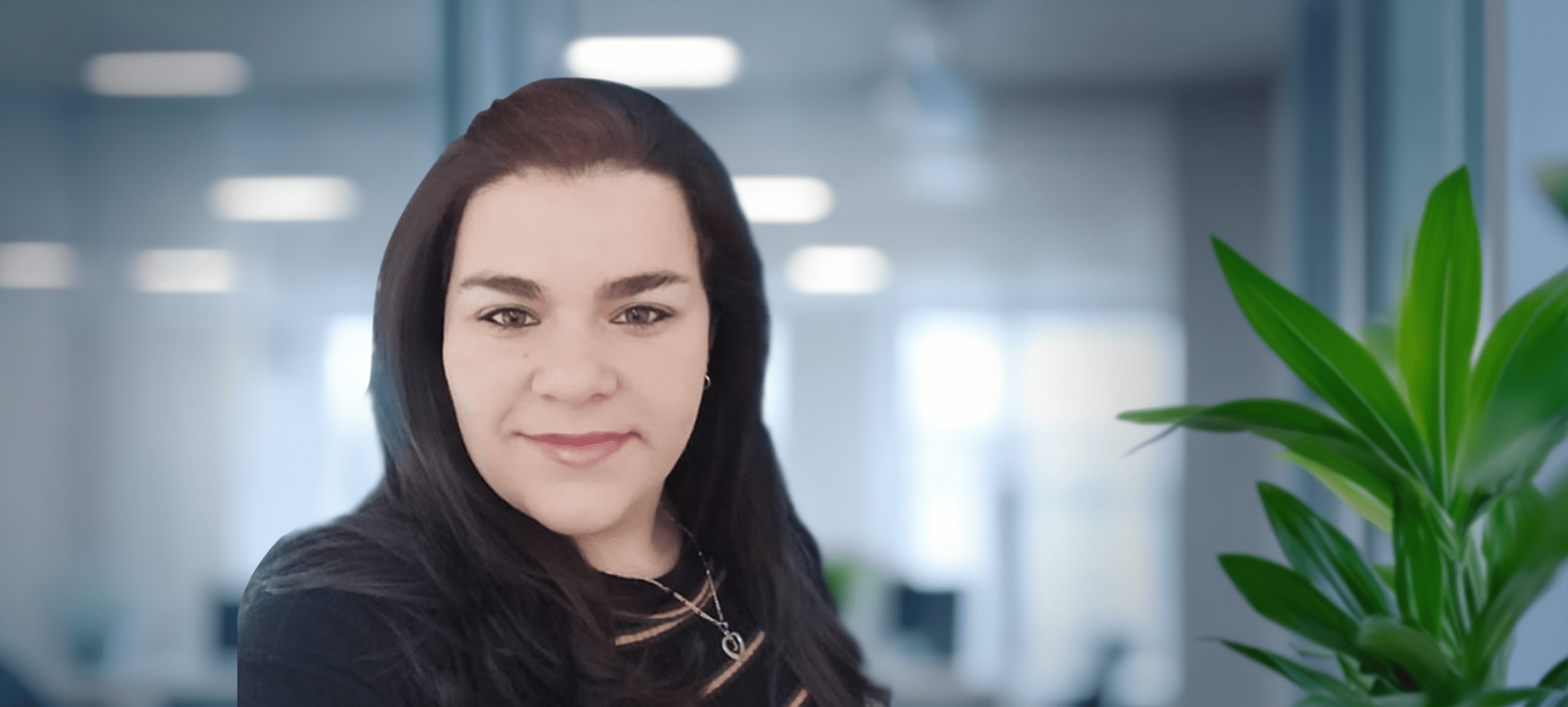 Adherent (formerly Compliance & Risks). Headshot of a woman with dark hair and a slight smile, wearing a dark top and necklace, with a blurred office background and a green plant.