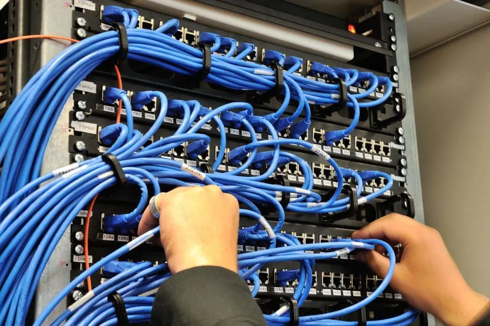 Adherent (formerly Compliance & Risks). Close-up of hands working with numerous blue Ethernet cables plugged into a server rack, showing organized cable management.