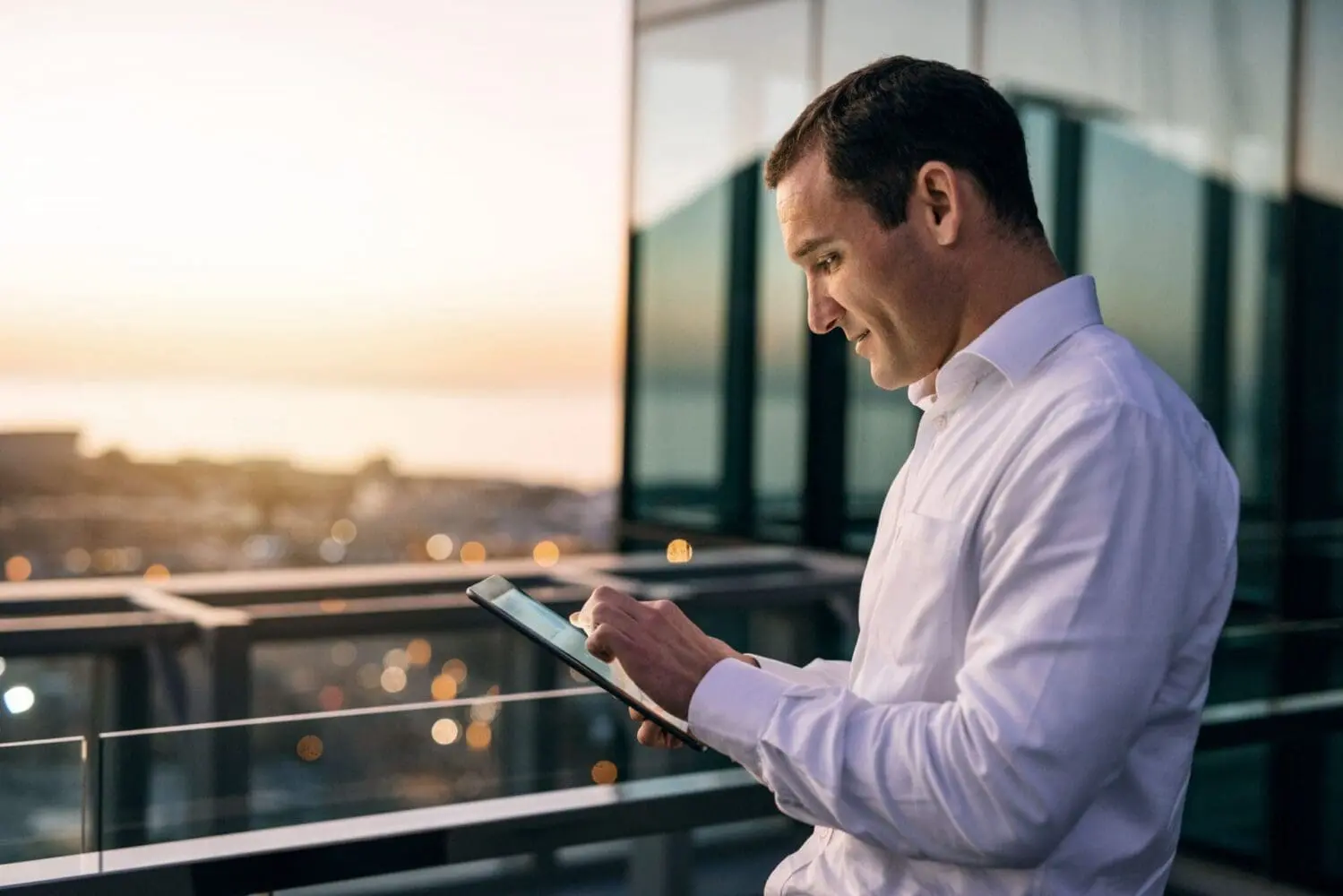 Adherent (formerly Compliance & Risks). A man in a white shirt uses a tablet on a balcony overlooking a city skyline at sunset.