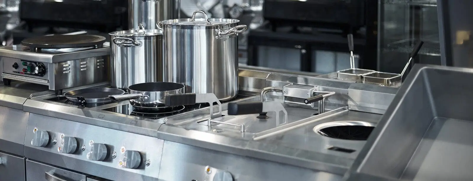 Adherent (formerly Compliance & Risks). A clean, modern commercial kitchen counter made of stainless steel, featuring a crepe maker, large stock pots, a saucepan, a deep fryer with baskets, and a sink.