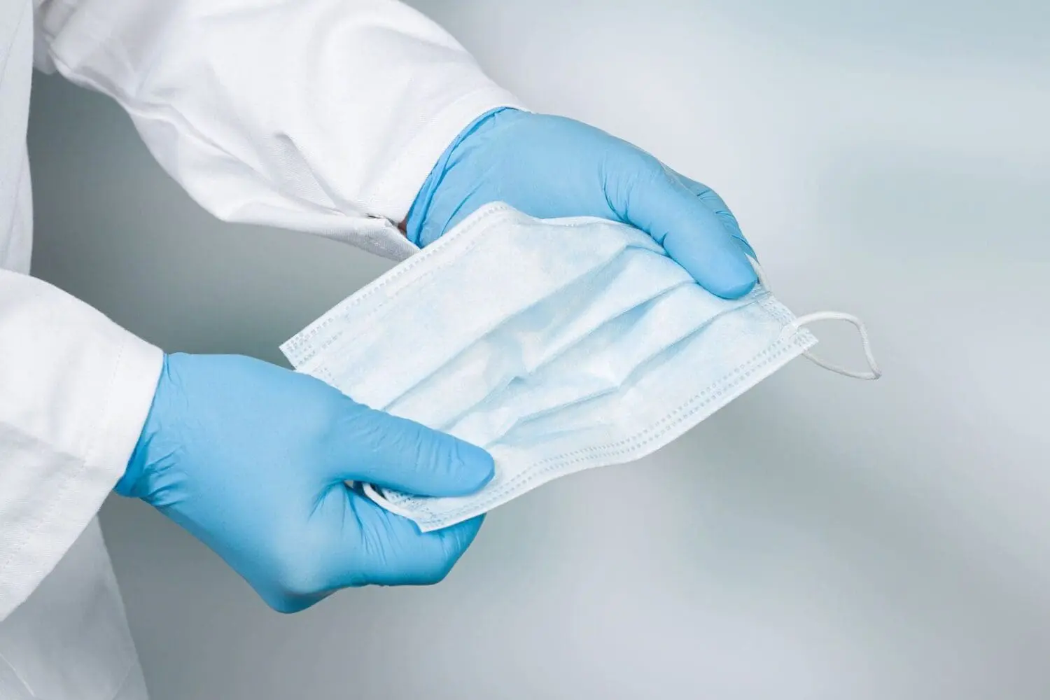 Adherent (formerly Compliance & Risks). Hands in blue medical gloves holding a light blue disposable surgical face mask.