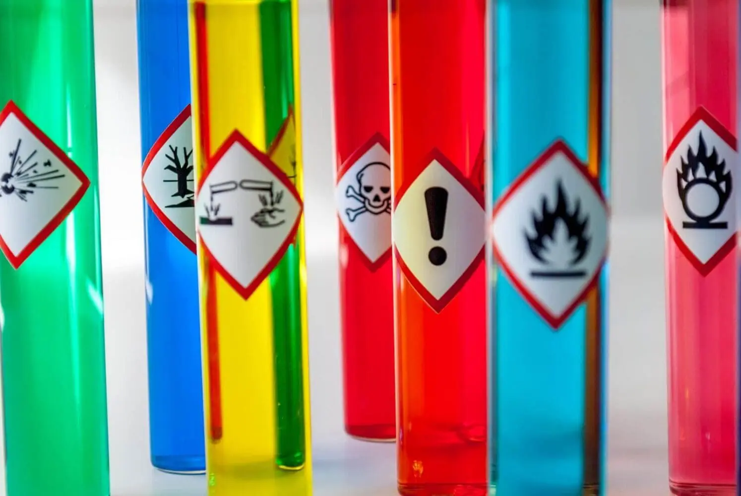 Adherent (formerly Compliance & Risks). Seven test tubes with colorful liquids and GHS hazard labels: exploding bomb, environmental hazard, corrosive, skull and crossbones, exclamation mark, flame, and flame over circle.
