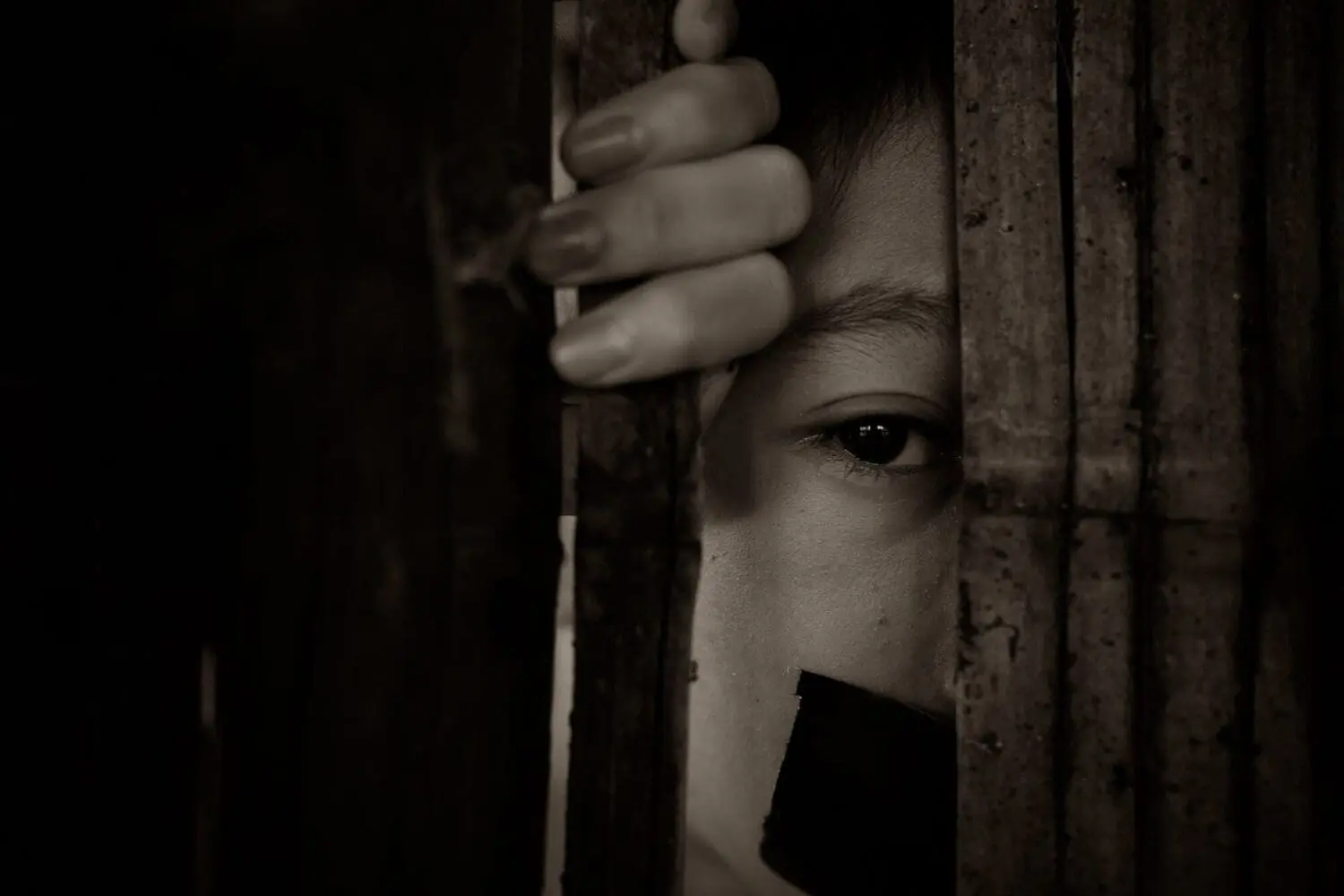 Adherent (formerly Compliance & Risks). Monochrome close-up of a person's eye and hand gripping wooden bars, with their mouth covered by dark tape.