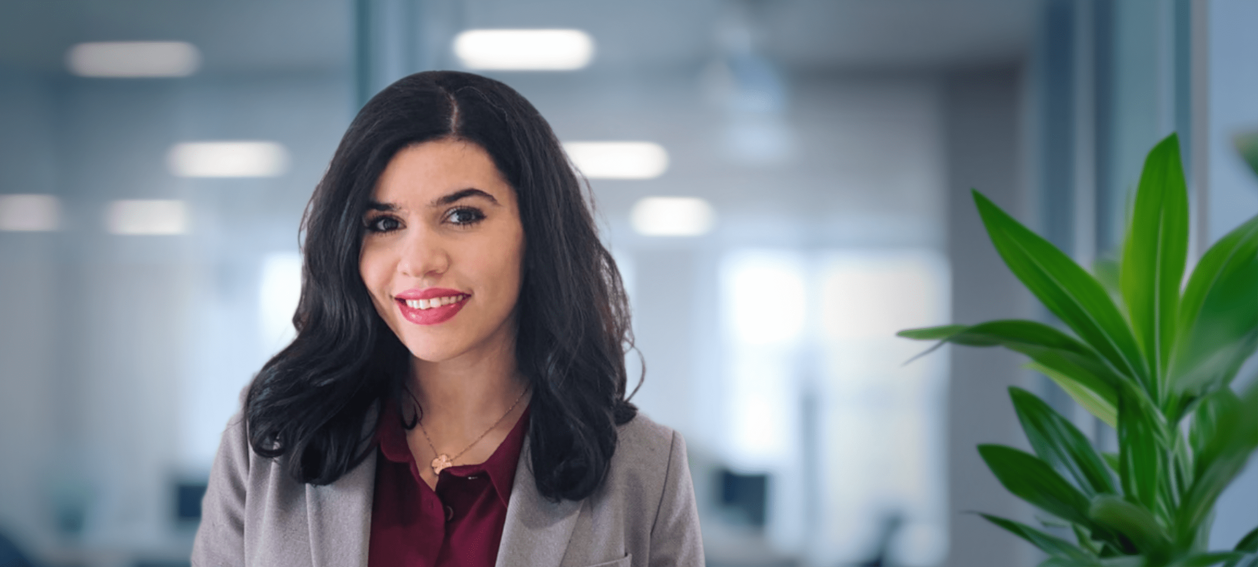 Adherent (formerly Compliance & Risks). A smiling woman with dark hair and red lipstick wears a gray blazer over a maroon shirt in an office.
