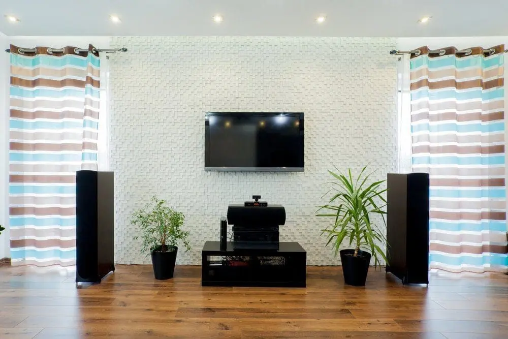 Adherent (formerly Compliance & Risks). A living room with a wall-mounted TV, black media console, two tall speakers, and potted plants on hardwood floor. Striped curtains frame the setup.