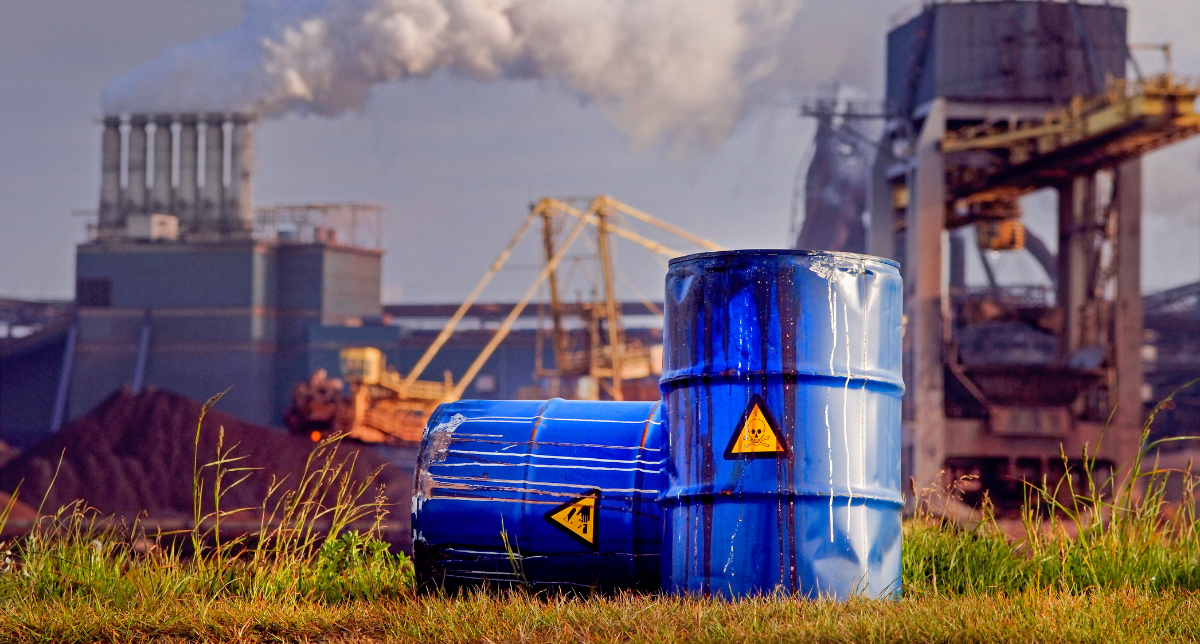 Adherent (formerly Compliance & Risks). Two blue industrial barrels with hazard symbols, stained with dark liquid, in front of a large factory emitting smoke.