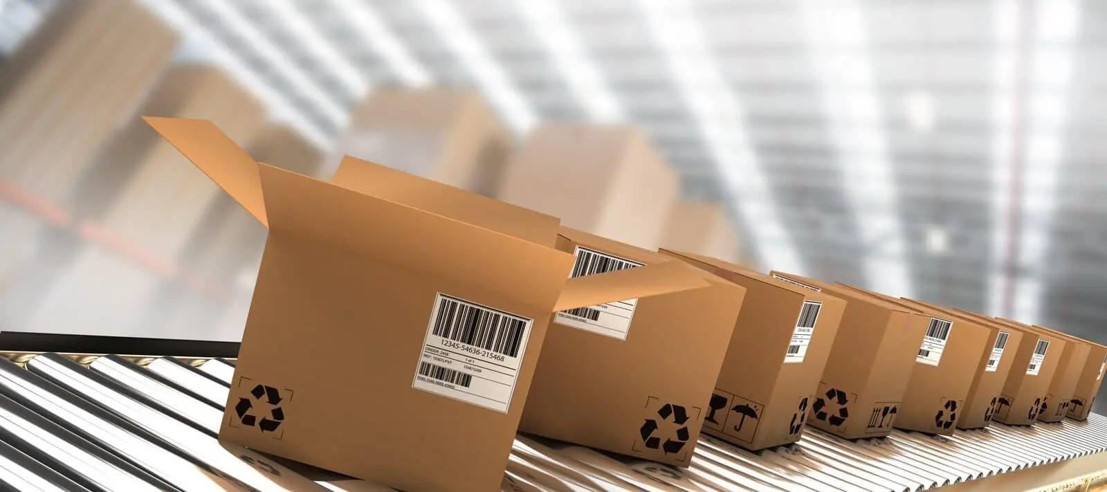 Brown cardboard boxes with barcodes and recycling symbols on a silver roller conveyor belt in a blurred warehouse.