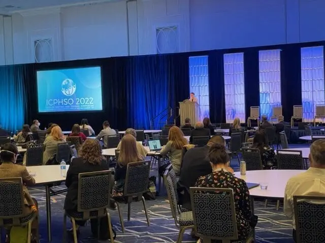 Adherent (formerly Compliance & Risks). A conference hall with attendees seated at tables facing a stage where a speaker is presenting in front of a screen displaying "ICPHSO 2022".