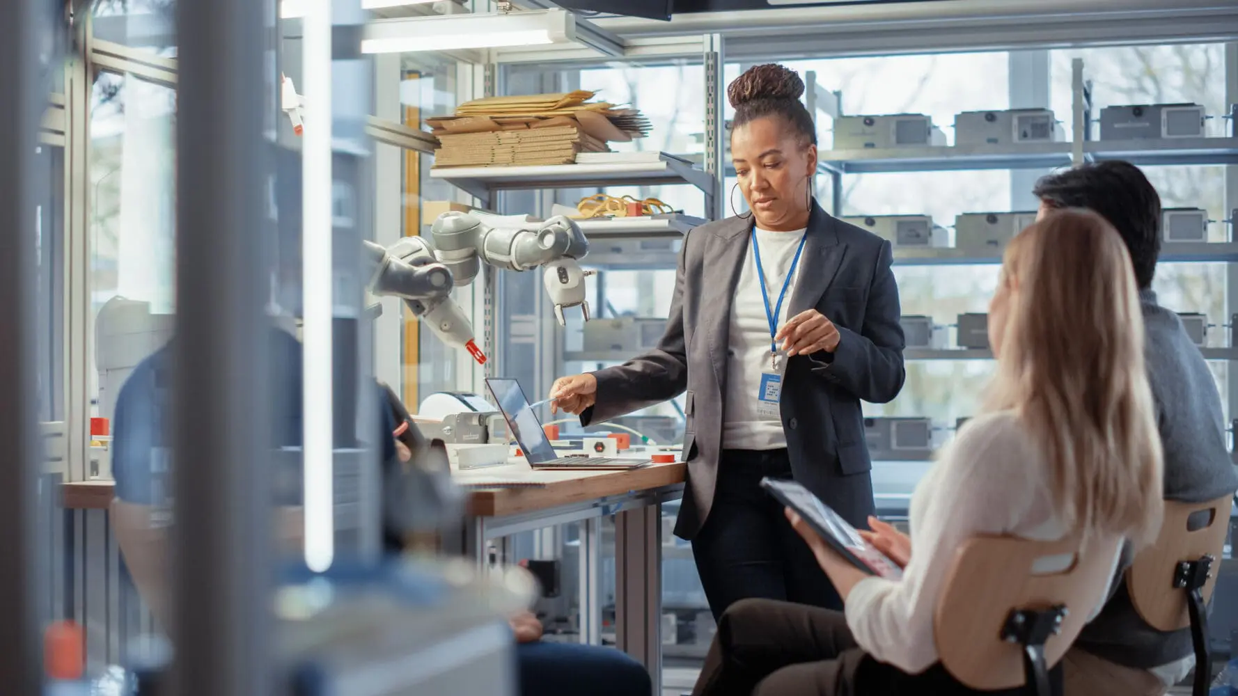 Adherent (formerly Compliance & Risks). An African American woman in a blazer points at a laptop on a workbench next to a robotic arm, while two colleagues observe in a high-tech lab.
