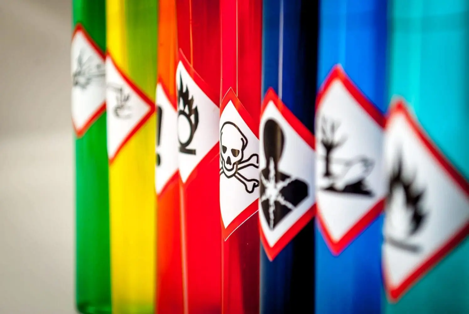 Adherent (formerly Compliance & Risks). A row of colorful bottles (green, yellow, orange, red, dark blue, light blue), each featuring a white diamond-shaped GHS hazard label. Visible pictograms include gas cylinder, exclamation mark, flame, skull and crossbones, human silhouette with starburst, and dead tree and fish.