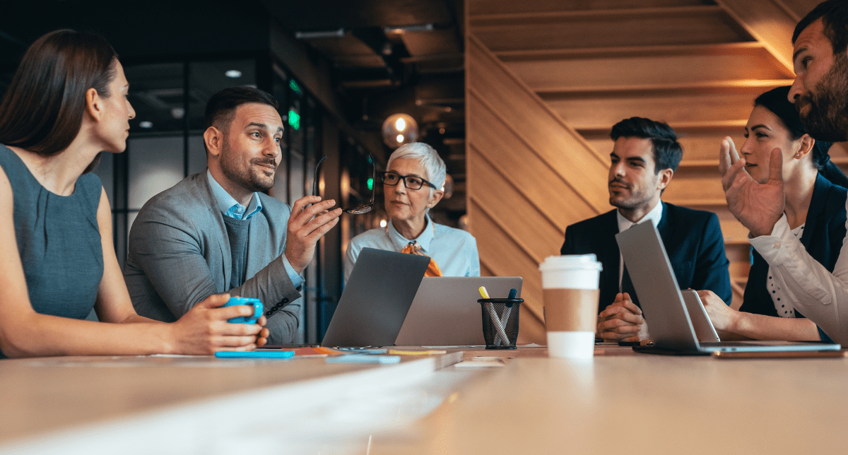 Adherent (formerly Compliance & Risks). Six diverse professionals, three men and three women, in a modern office meeting, some with laptops, engaged in discussion.