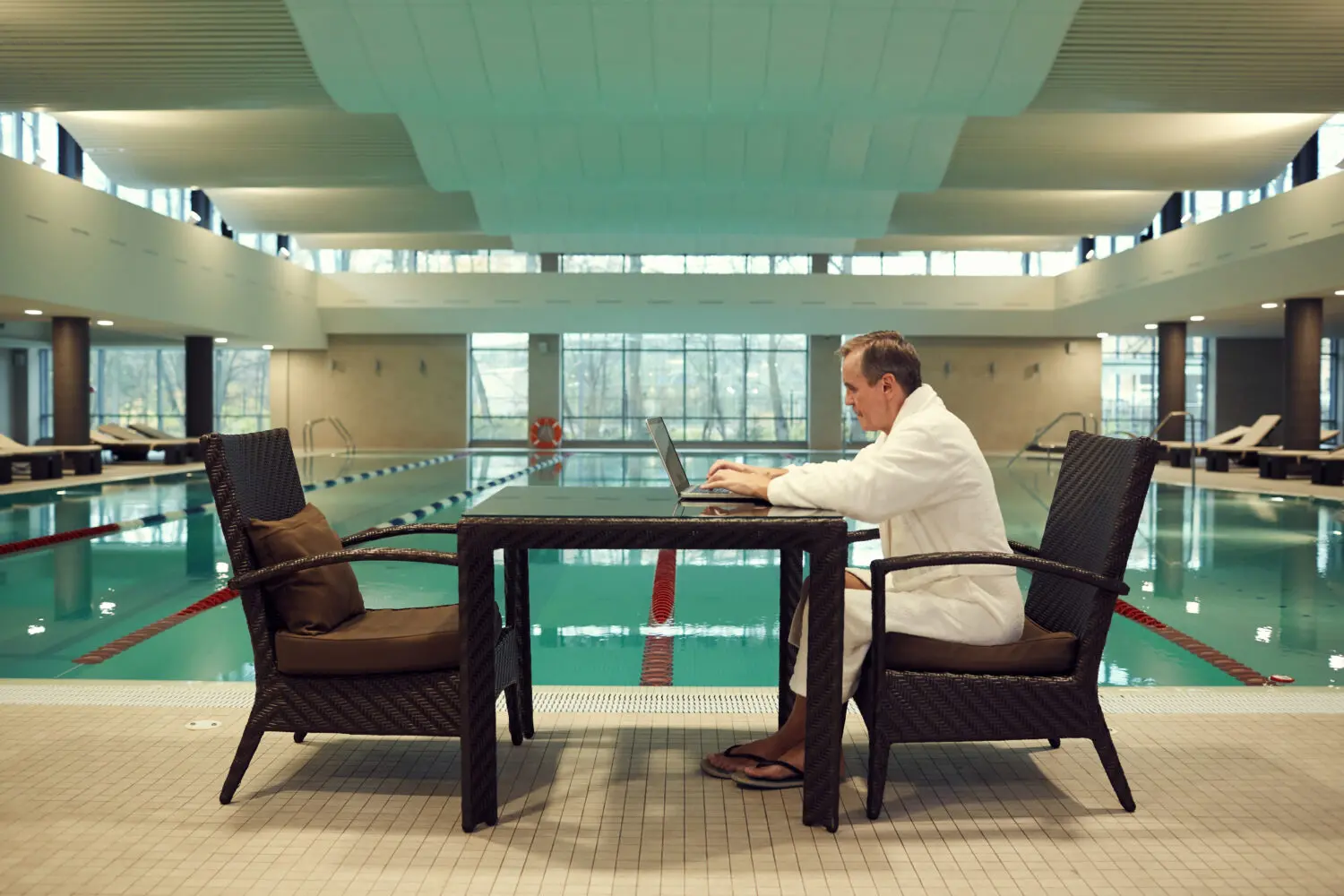 Adherent (formerly Compliance & Risks). A man in a white bathrobe sits at a wicker table, typing on a laptop next to a large indoor swimming pool.