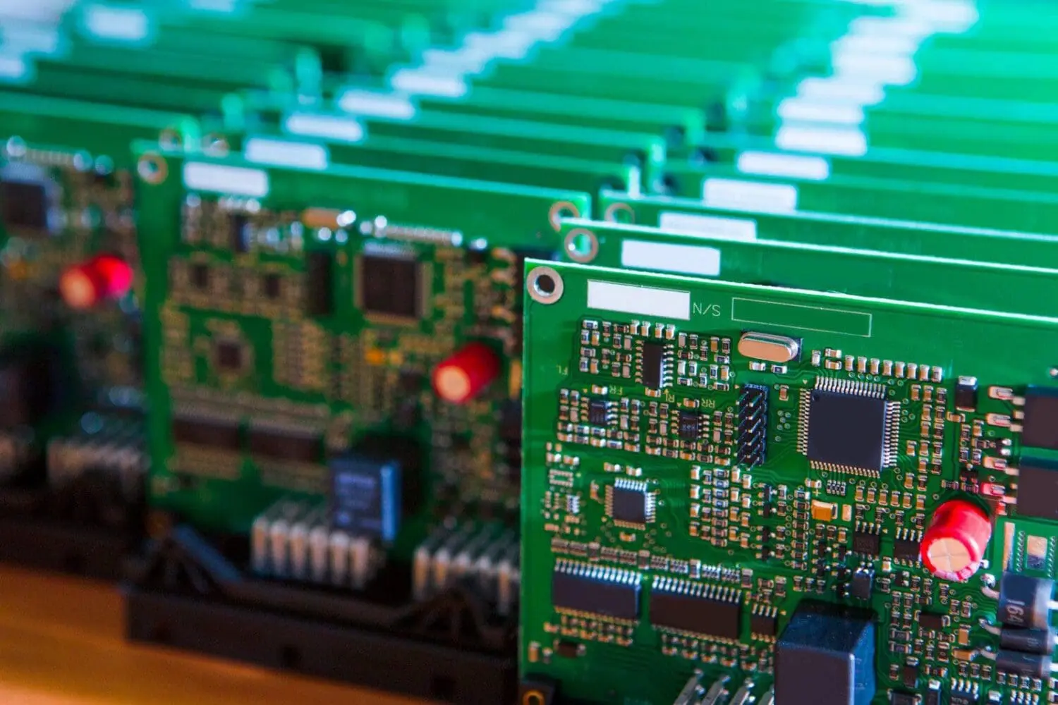 Adherent (formerly Compliance & Risks). Close-up of several green circuit boards with various electronic components, including black IC chips, red capacitors, and metal oscillators.