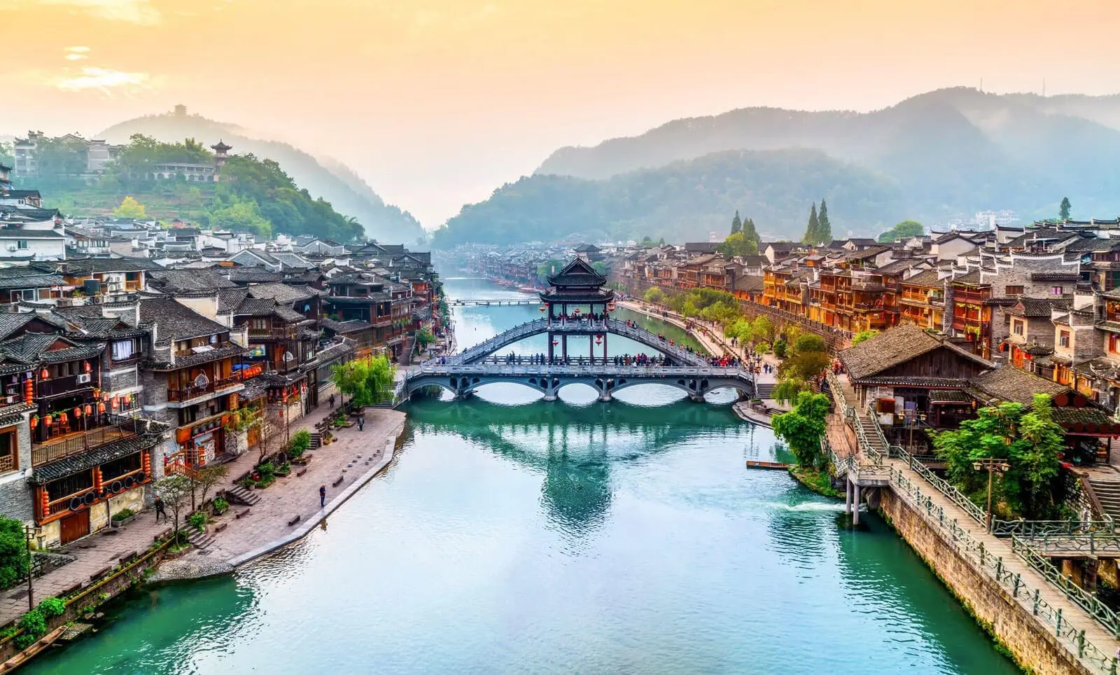 Adherent (formerly Compliance & Risks). Panoramic view of Fenghuang Ancient Town with a river, a large arched bridge with a pavilion, traditional buildings lining the banks, and hazy mountains in the background under a warm, golden sky.