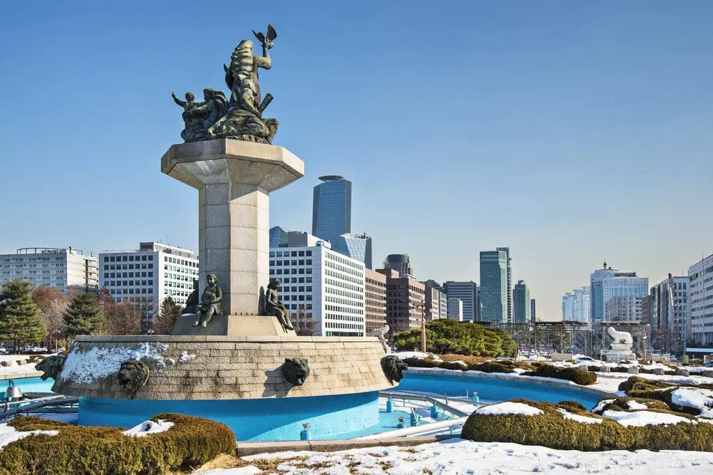 Adherent (formerly Compliance & Risks). A large bronze statue on a stone pedestal with a blue fountain in front, surrounded by snow and green bushes, with a city skyline in the background under a clear sky.