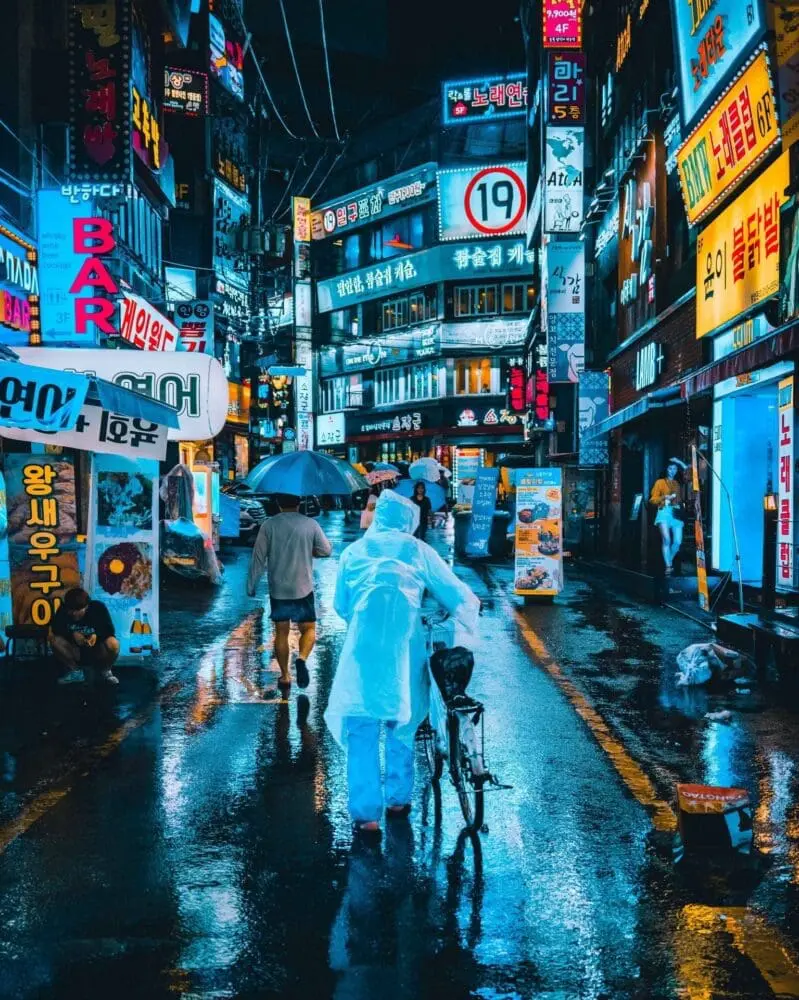 Adherent (formerly Compliance & Risks). A busy city street at night, wet from rain, with numerous brightly lit neon signs in Korean. People walk with umbrellas and a person pushes a bicycle, their figures reflected in the wet ground.