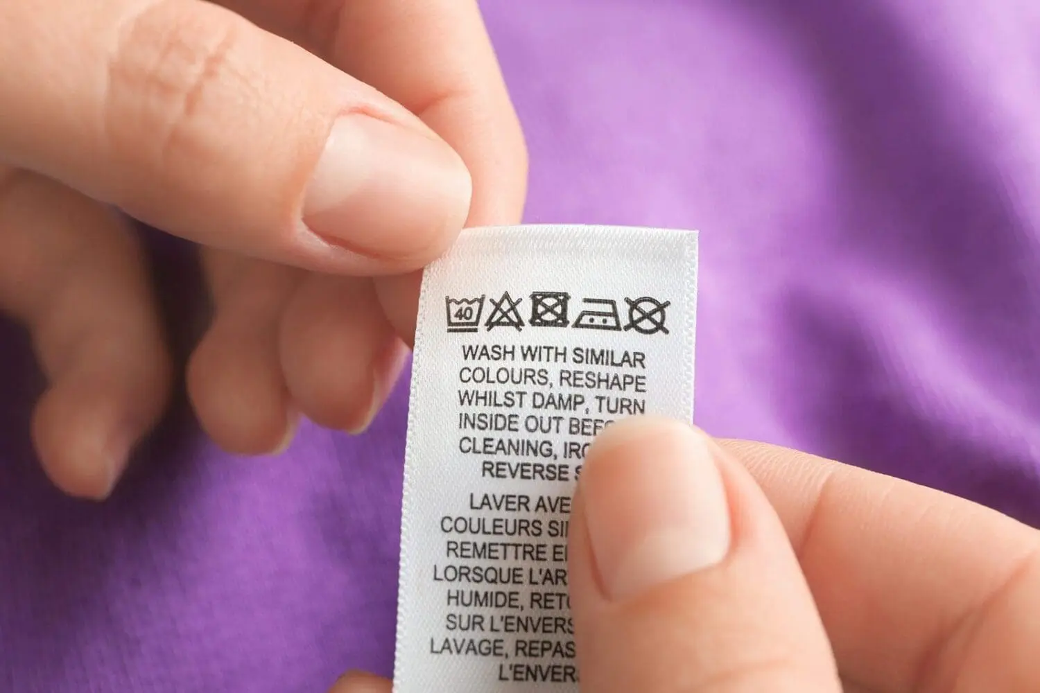 Adherent (formerly Compliance & Risks). Close-up of hands holding a white clothing care label with laundry symbols and text against a blurred purple background.