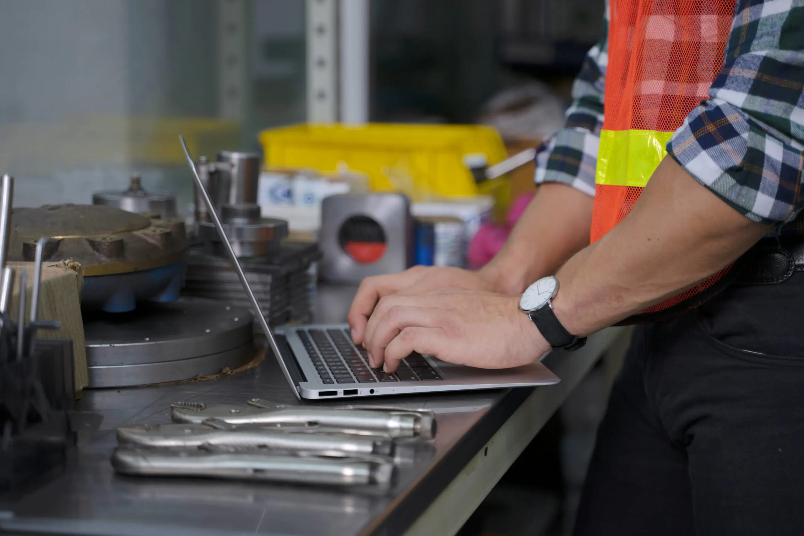 Close-up of a person in a plaid shirt and orange safety vest typing on a silver laptop on a metal workbench, surrounded by tools and industrial equipment.