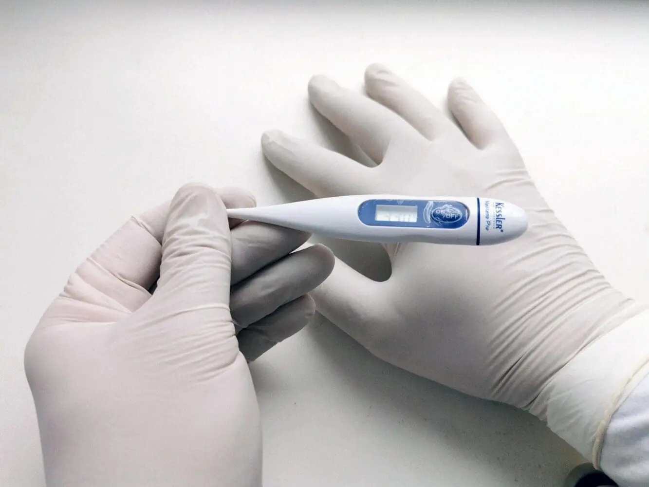 Adherent (formerly Compliance & Risks). Two gloved hands hold a white and blue digital thermometer displaying '33.0' on its screen.