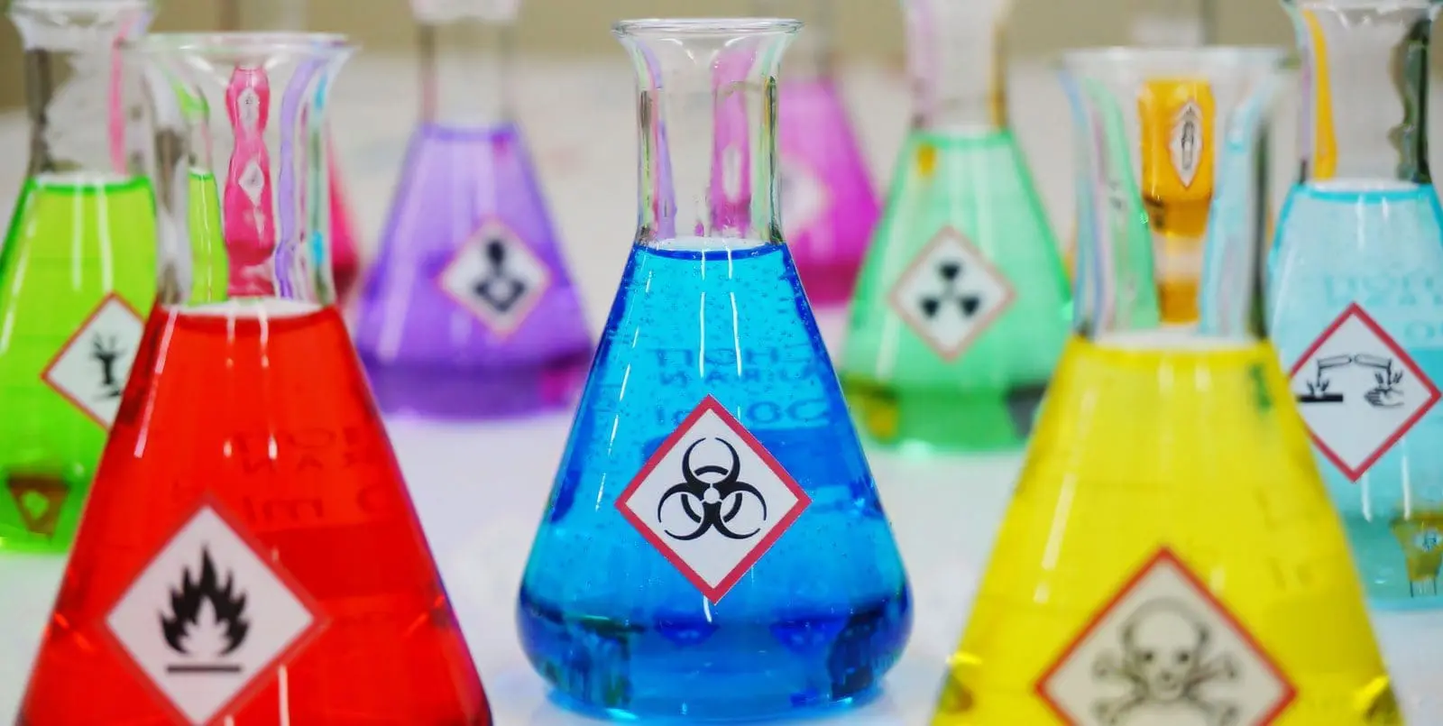 Adherent (formerly Compliance & Risks). Multiple colorful Erlenmeyer flasks, each with a different colored liquid and a hazard symbol. The central blue flask has a biohazard symbol, the red flask has a flame, and the yellow flask has a skull and crossbones.