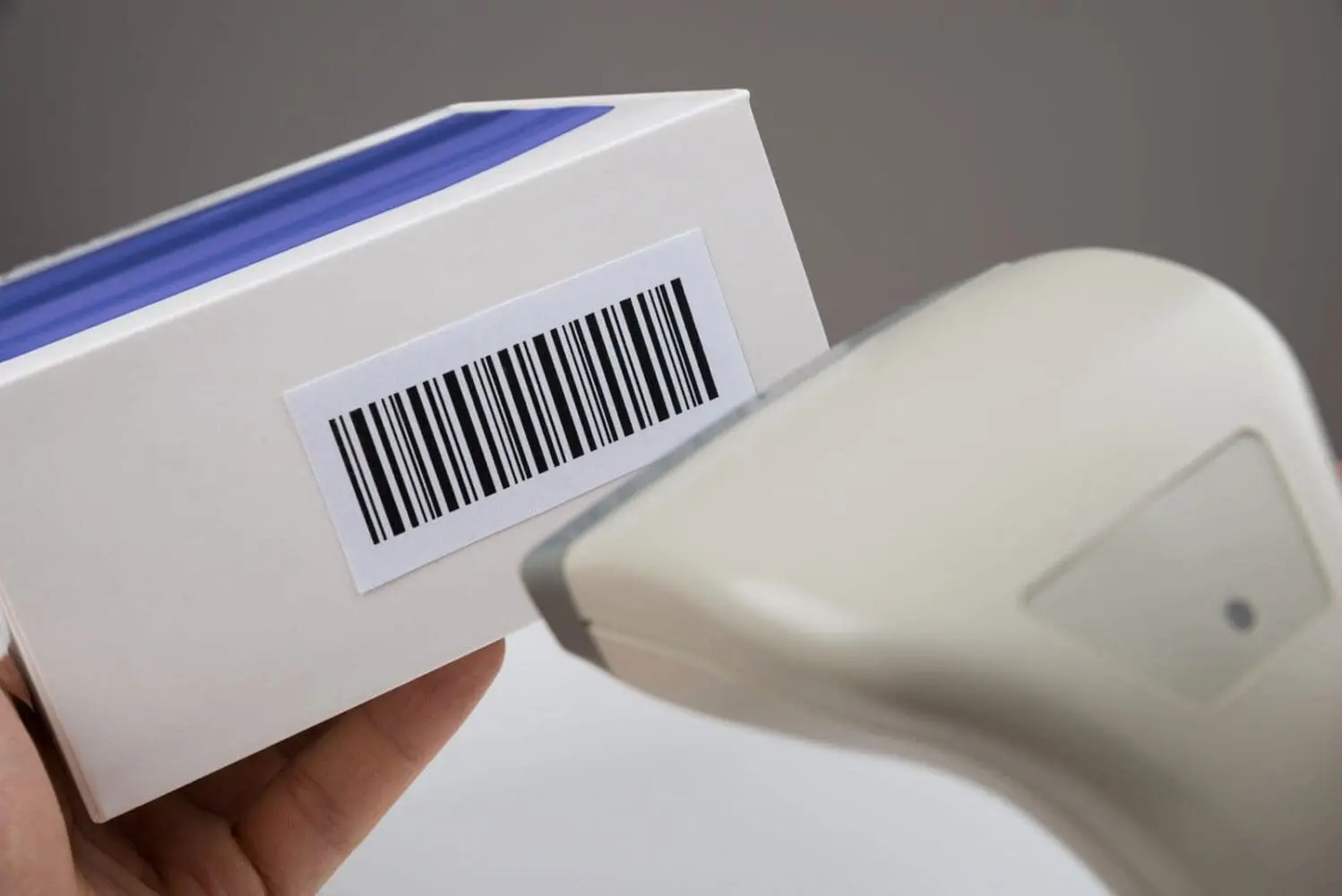 Adherent (formerly Compliance & Risks). A hand holds a white box with a barcode, being scanned by a light-colored barcode scanner.