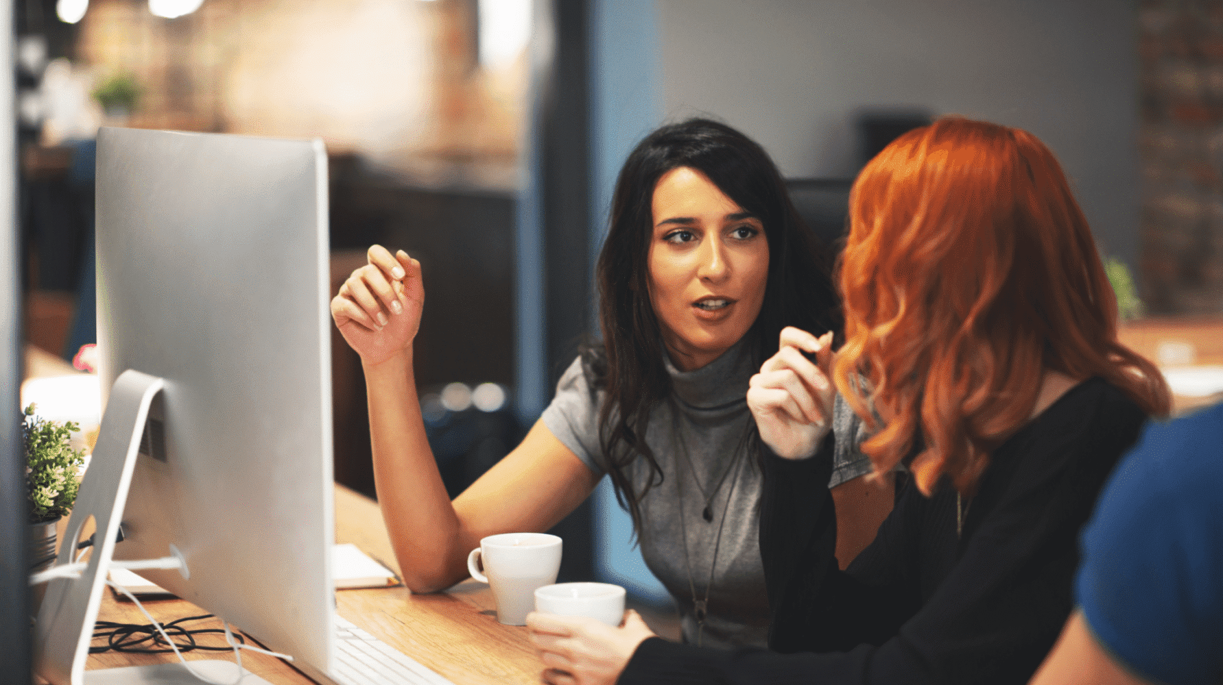 Adherent (formerly Compliance & Risks). Two women at a desk, one with dark hair speaking and gesturing, the other with red hair listening and holding a mug, with a computer monitor between them.