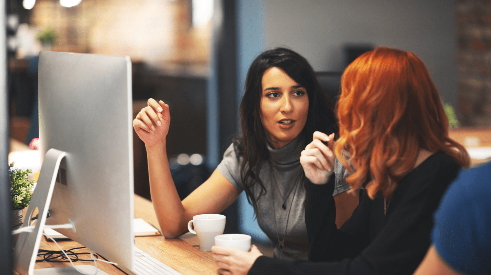 Two women at a desk, one with dark hair speaking and gesturing, the other with red hair listening and holding a mug, with a computer monitor between them.