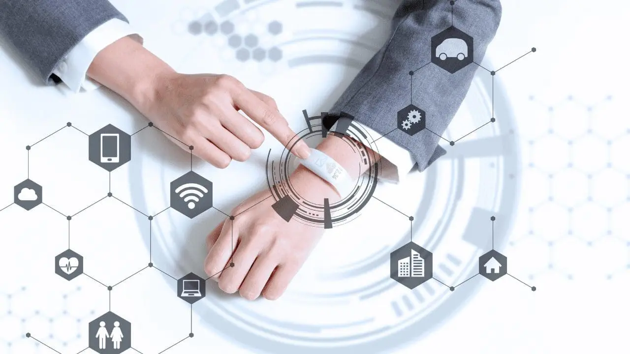 A person's hands interact with a white wearable device on their wrist, surrounded by a digital overlay of interconnected hexagonal icons representing cloud, smartphone, Wi-Fi, health, laptop, people, car, gears, building, and home.