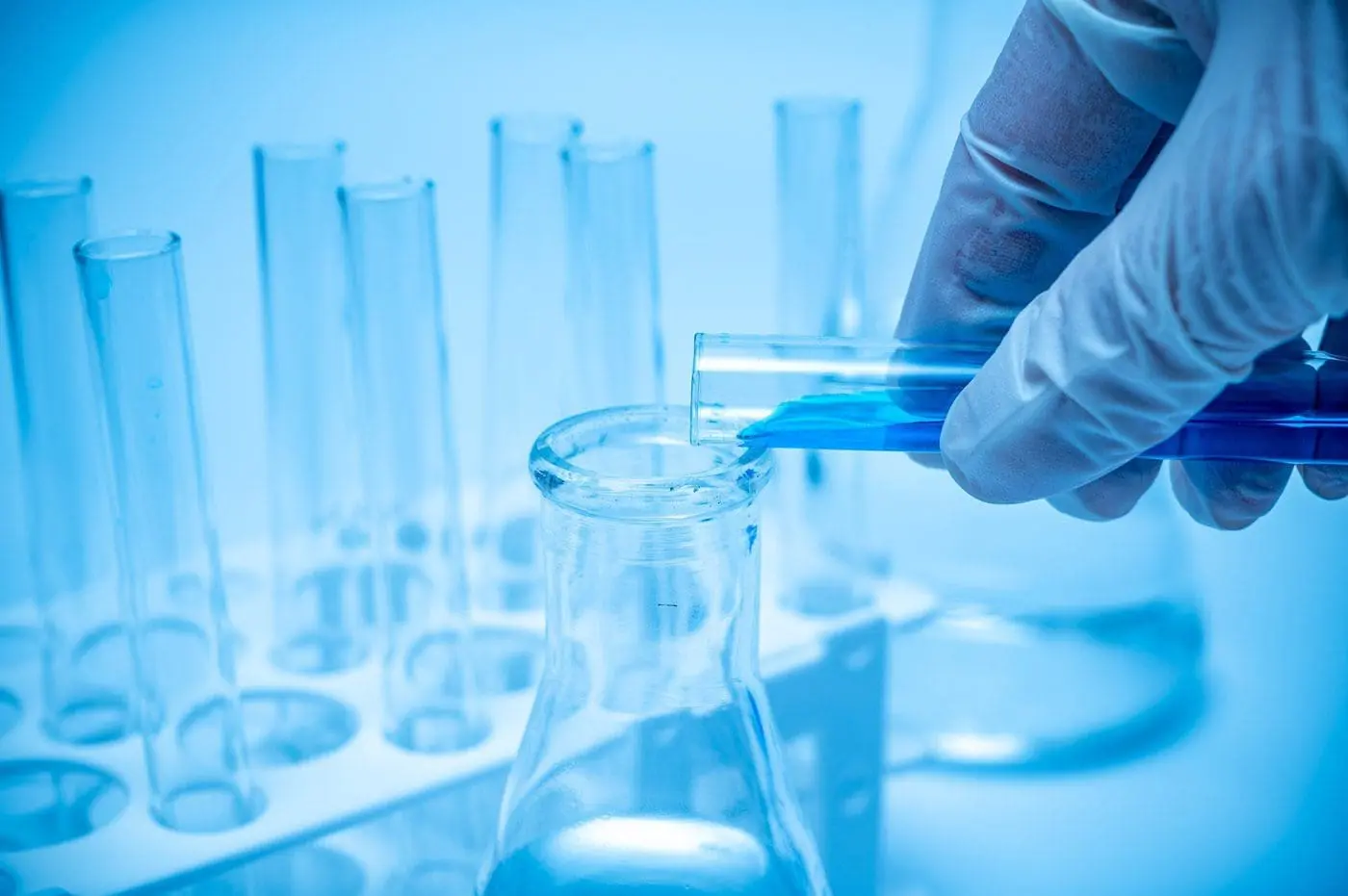 Adherent (formerly Compliance & Risks). A gloved hand pours blue liquid from a test tube into a flask in a lab setting with other test tubes.