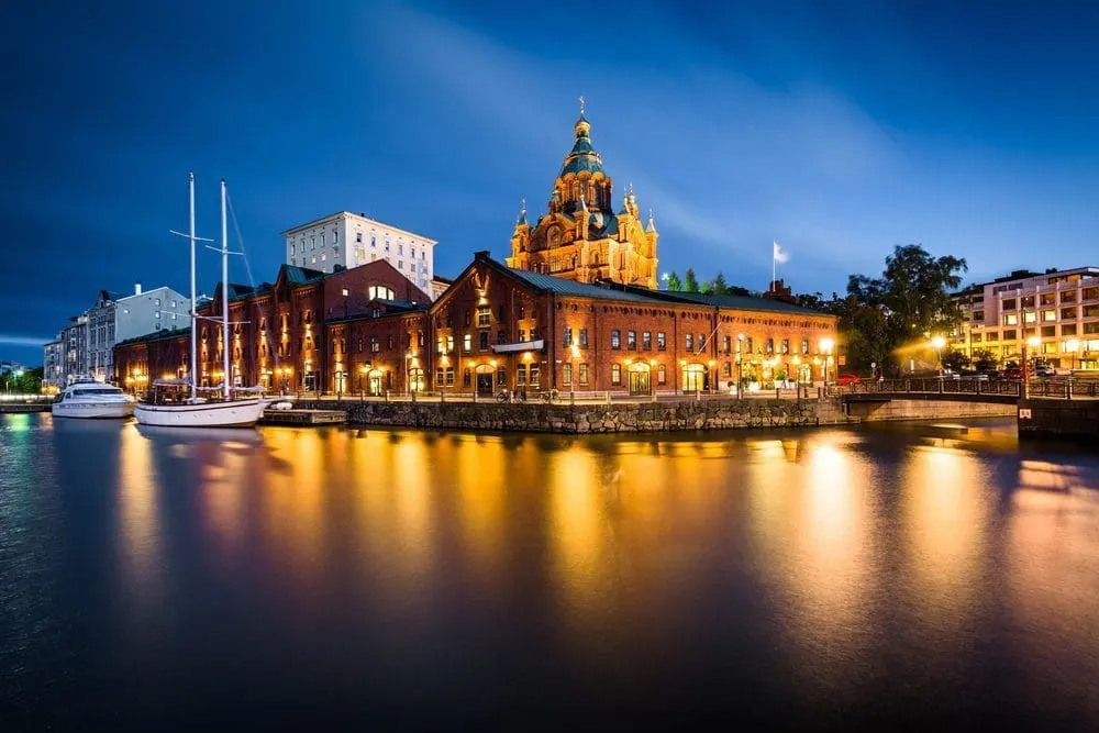 Adherent (formerly Compliance & Risks). Night view of Helsinki's waterfront with illuminated brick buildings, docked boats, and the brightly lit Uspenski Cathedral reflecting in the calm water.