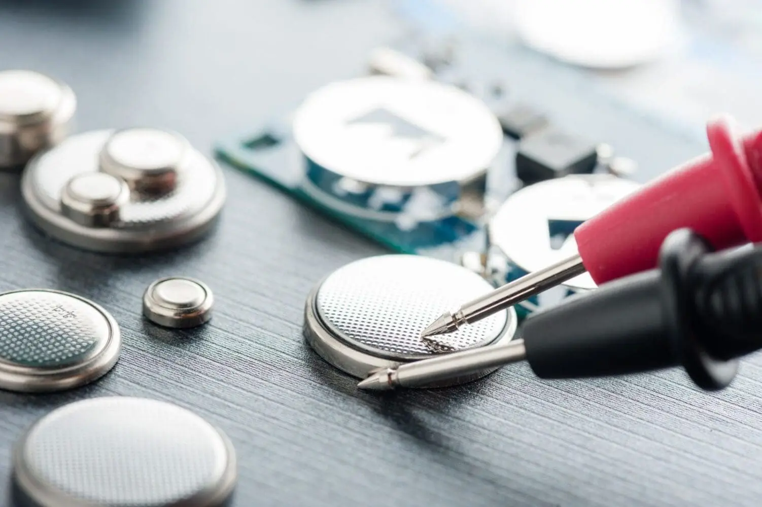 Adherent (formerly Compliance & Risks). Close-up of a red and a black multimeter probe touching a silver coin cell battery, with other coin cell batteries and a blurred circuit board in the background.