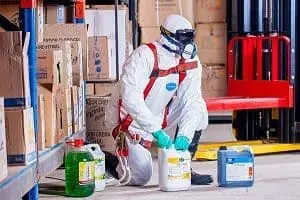 Adherent (formerly Compliance & Risks). Person in a white hazmat suit, respirator, and gloves kneeling next to various chemical containers in a warehouse.