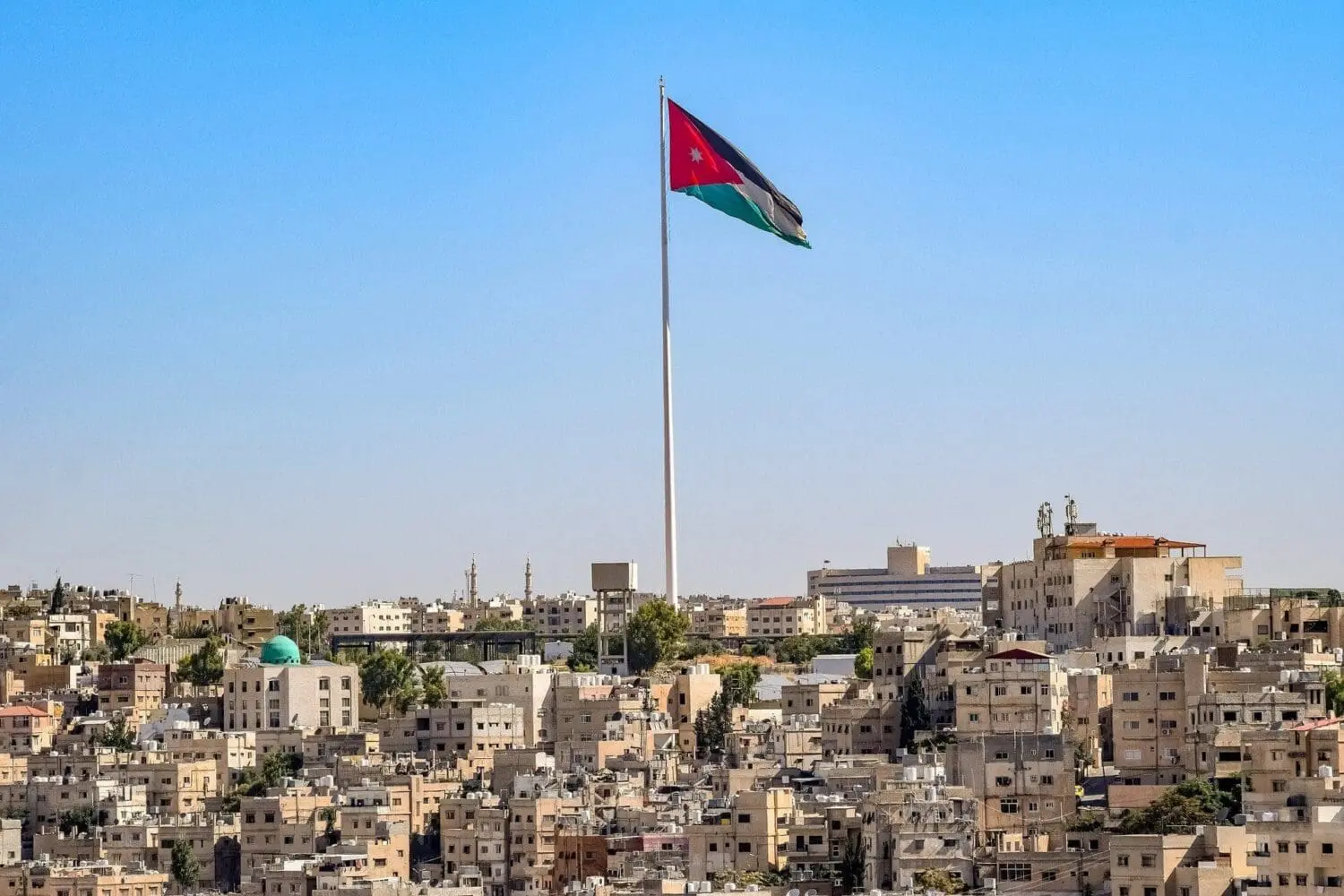 Adherent (formerly Compliance & Risks). The Jordanian flag on a very tall flagpole overlooking a dense city with light-colored buildings and a clear blue sky.