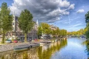 Adherent (formerly Compliance & Risks). A city canal lined with historic brick buildings, green trees, and moored boats under a partly cloudy blue sky.
