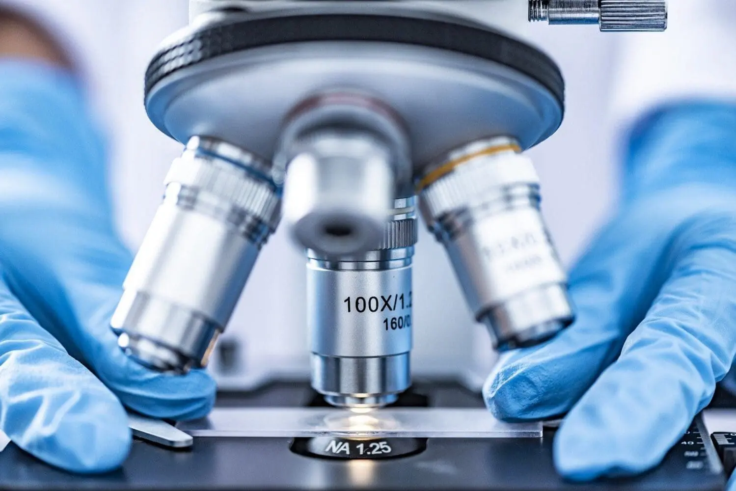 Adherent (formerly Compliance & Risks). Close-up of gloved hands operating a microscope with a slide under the 100x objective lens, illuminated from below.