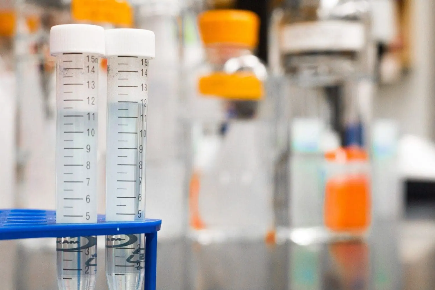 Adherent (formerly Compliance & Risks). Two clear, graduated centrifuge tubes with white caps, containing clear liquid, standing in a blue rack in a blurred laboratory setting.