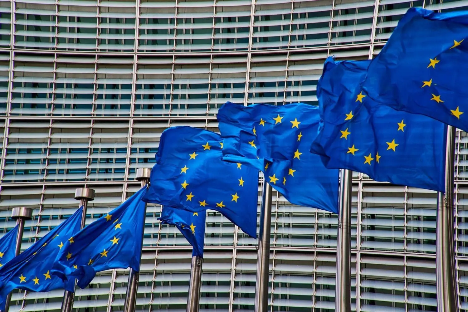 Several blue European Union flags with yellow stars wave in front of a modern building with horizontal louvers.