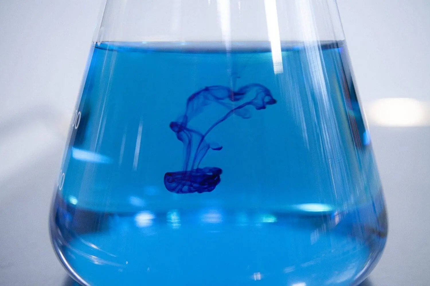 Adherent (formerly Compliance & Risks). A clear glass Erlenmeyer flask filled with bright blue liquid, with darker blue dye diffusing and swirling downwards into it.