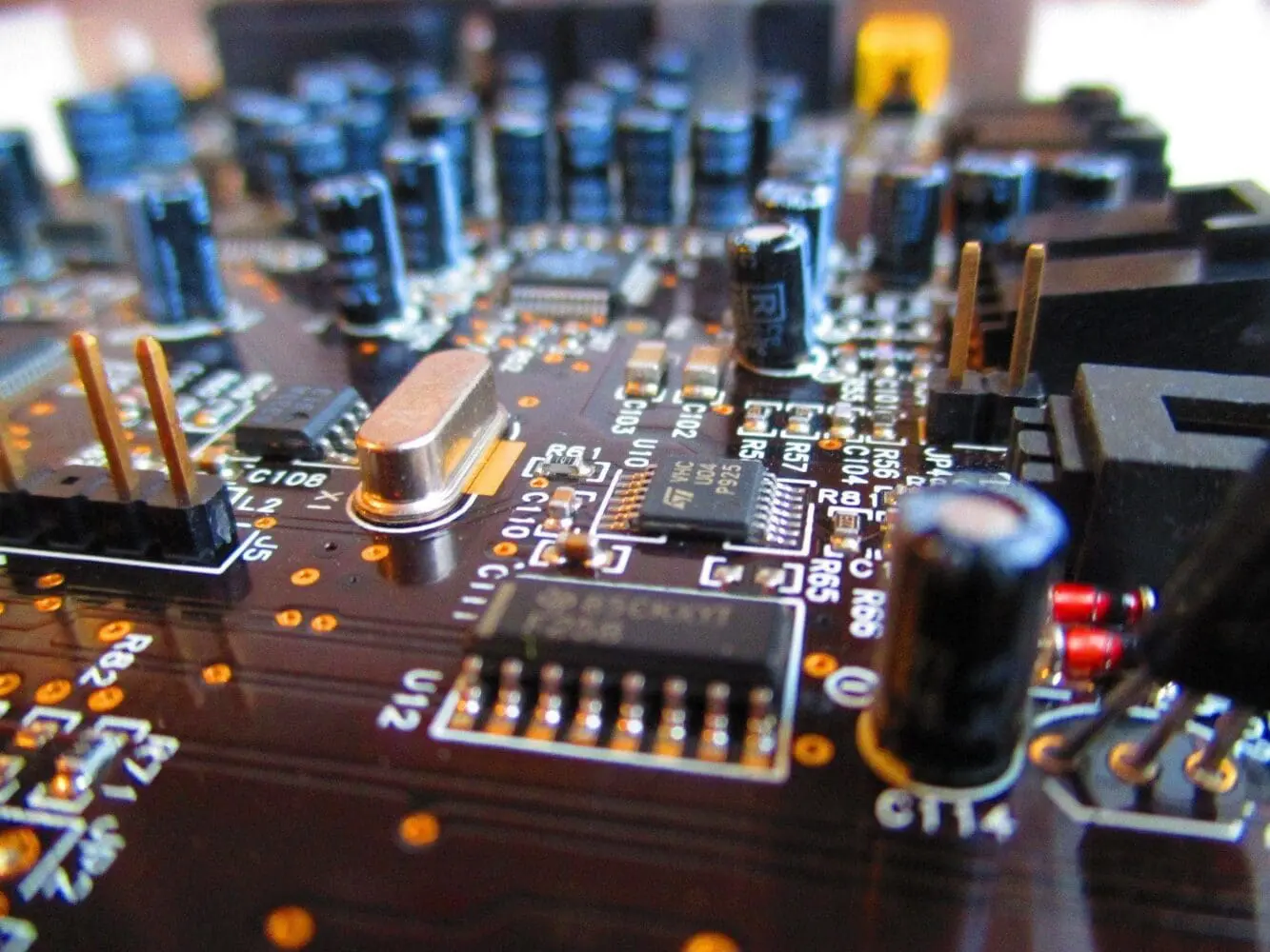 Close-up of a dark brown circuit board with numerous electronic components, including blue and black capacitors, black integrated circuits, gold connector pins, and a silver crystal oscillator.