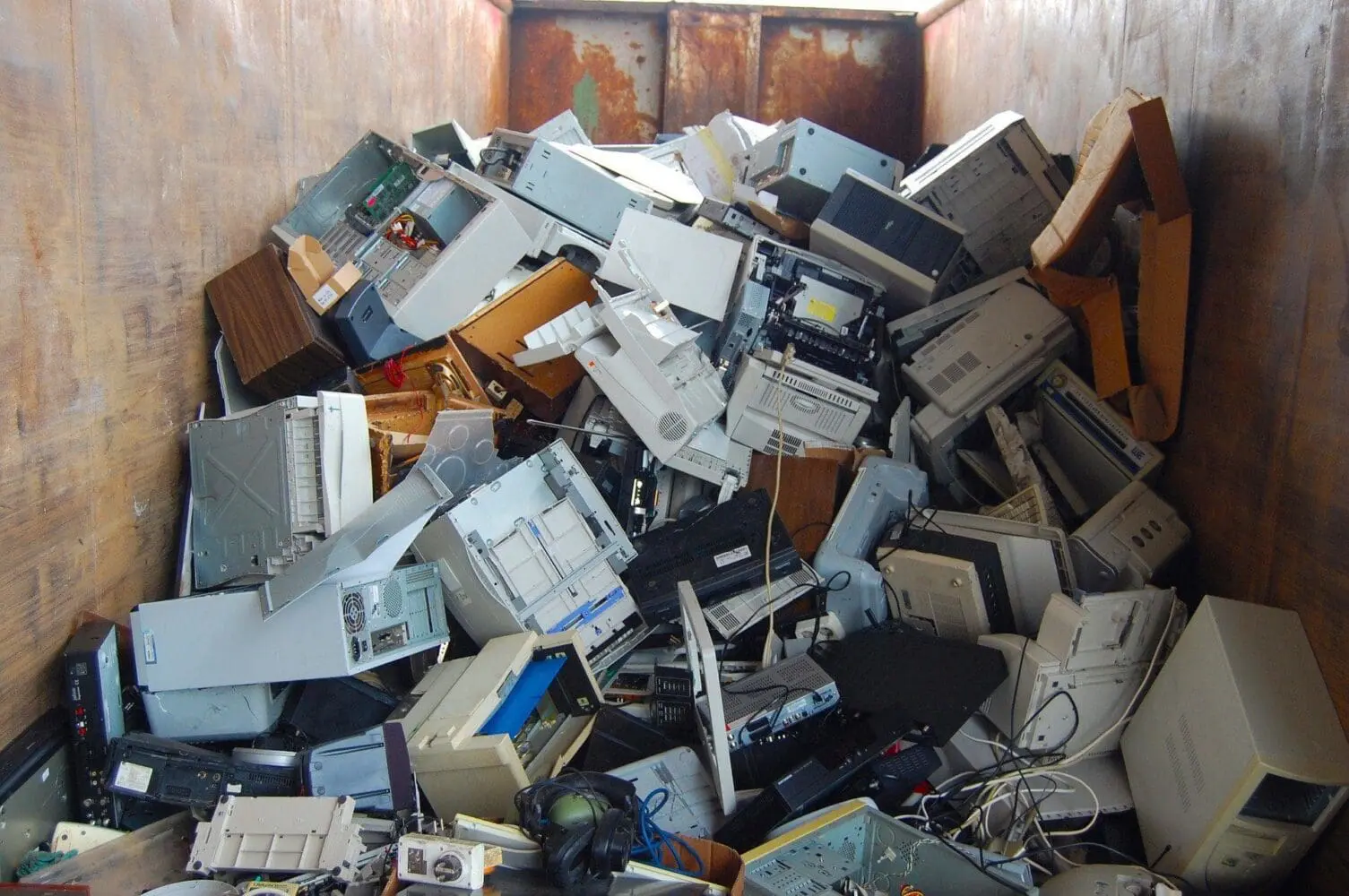 A large pile of electronic waste, including computer towers, printers, and monitors, inside a rusty metal container.