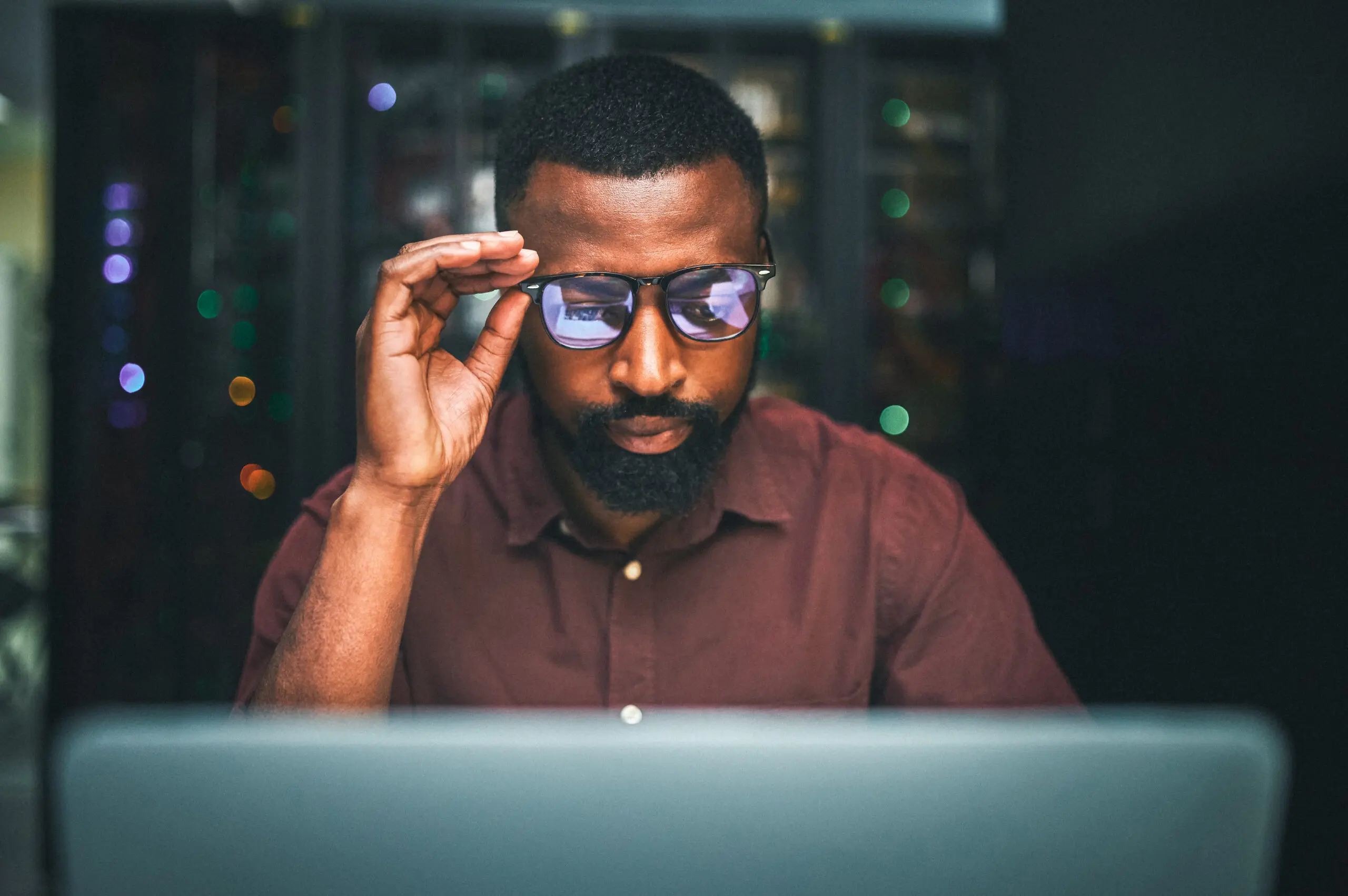 Adherent (formerly Compliance & Risks). A man in a maroon shirt adjusts his glasses while looking at a laptop screen, with blurred server lights in the background.