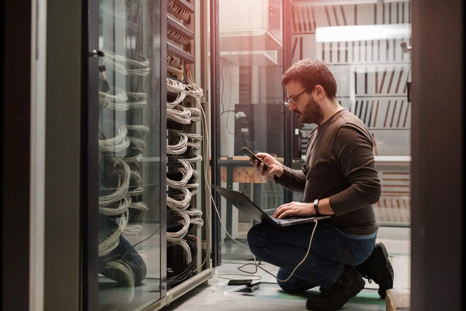 Adherent (formerly Compliance & Risks). A man kneels in front of an open server rack, using a laptop and smartphone to work on the network cables and equipment.
