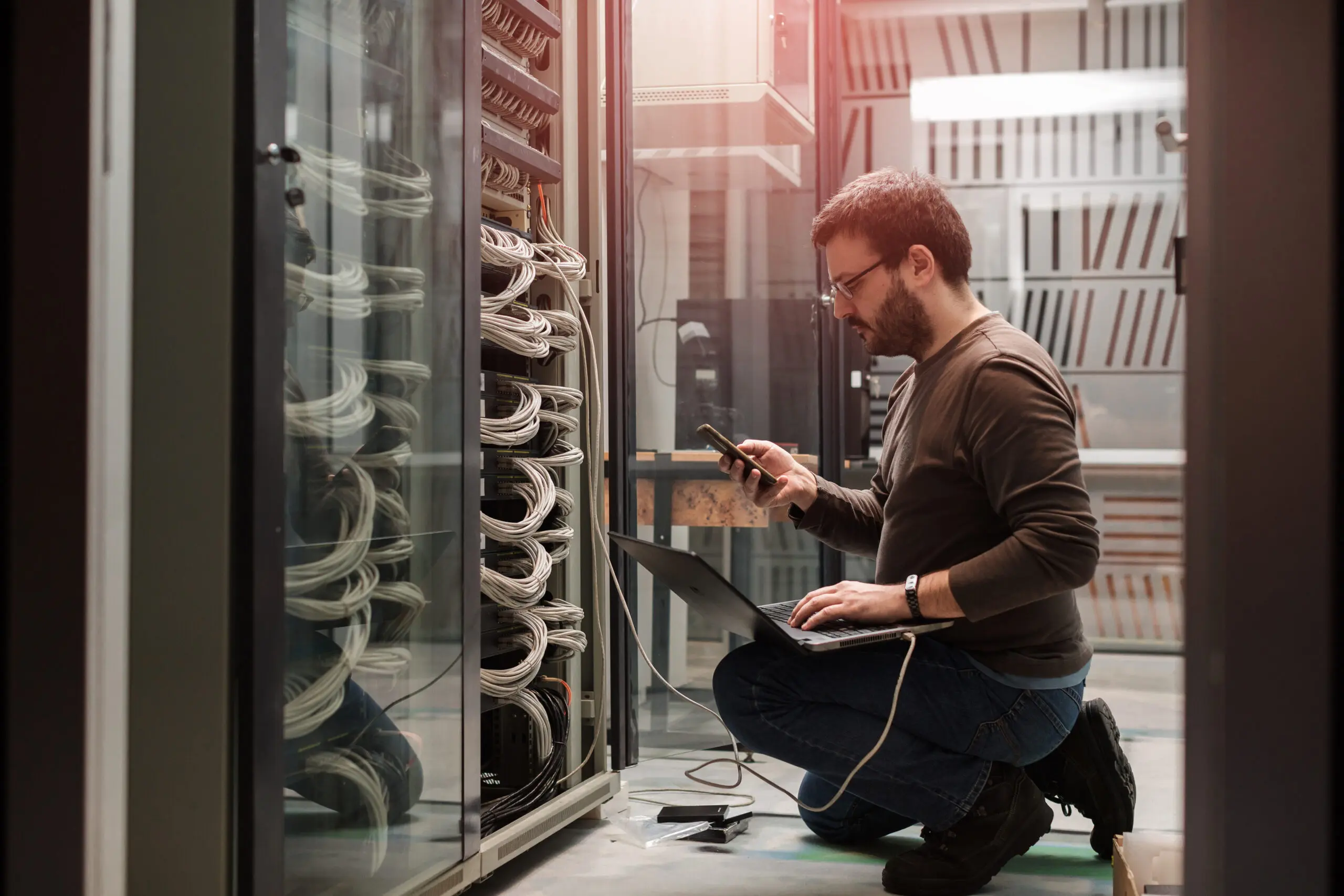 A man kneels in front of an open server rack, using a laptop and smartphone to work on the network cables and equipment.